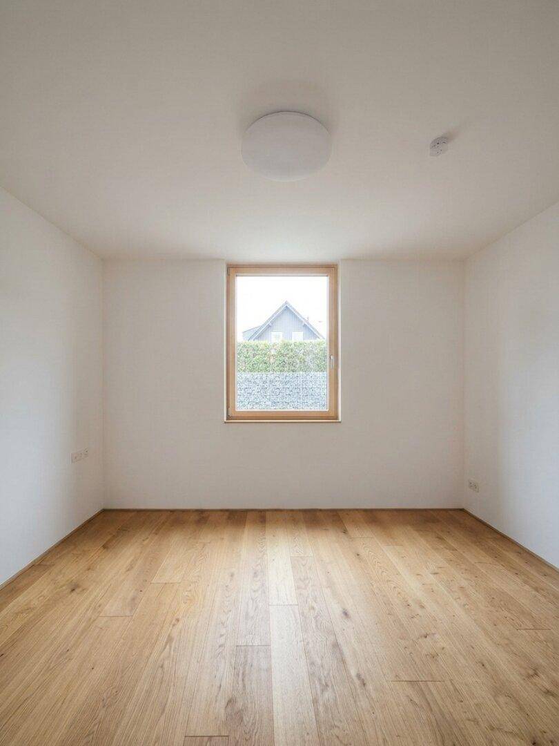 Leerer Raum mit hellem Holzboden und einem Fenster, das einen Blick ins Grüne bietet.