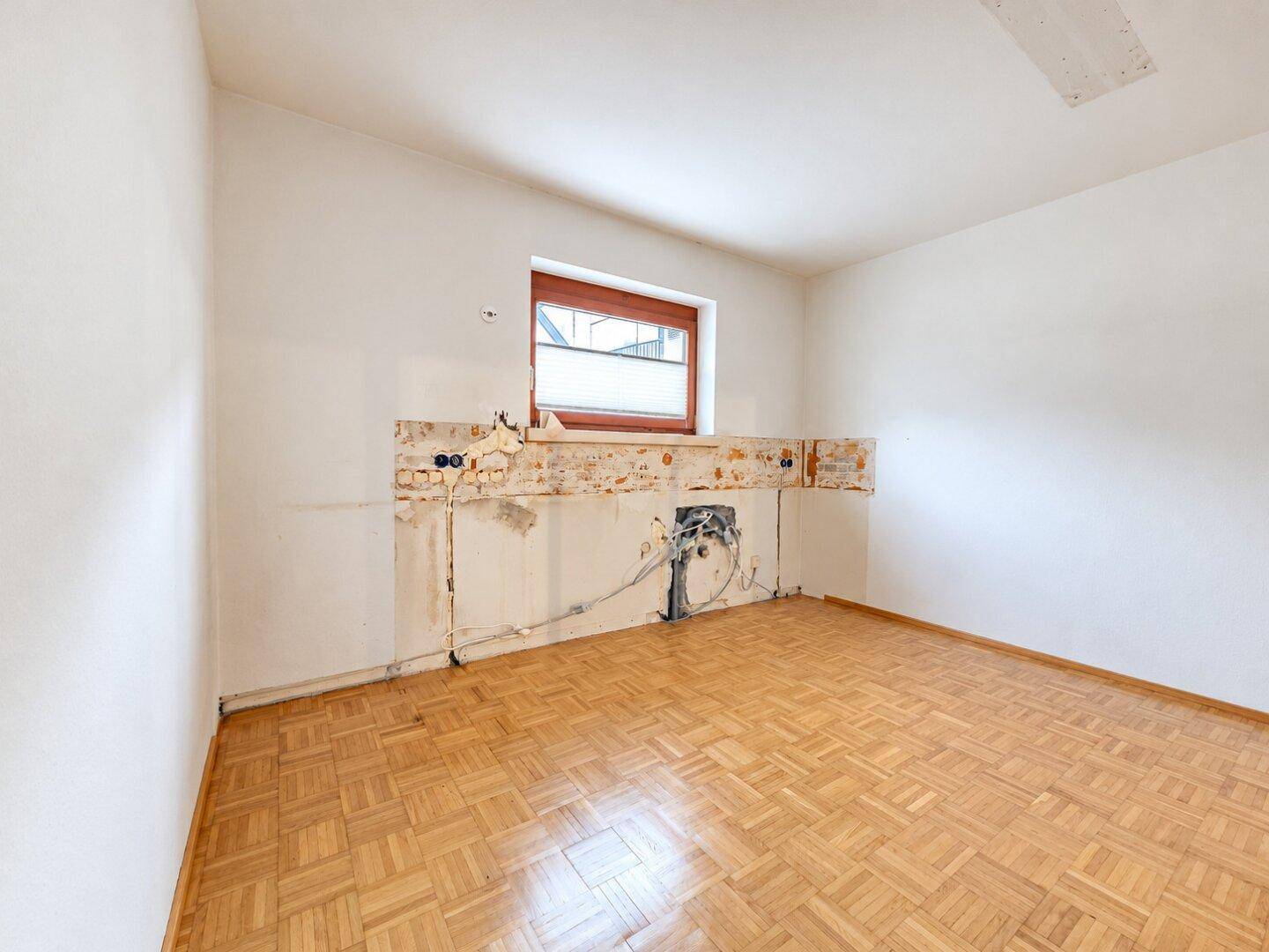 Leerer Küchenraum mit Parkettboden, Fenster und vorbereiteten Anschlüssen für eine Einbauküche.