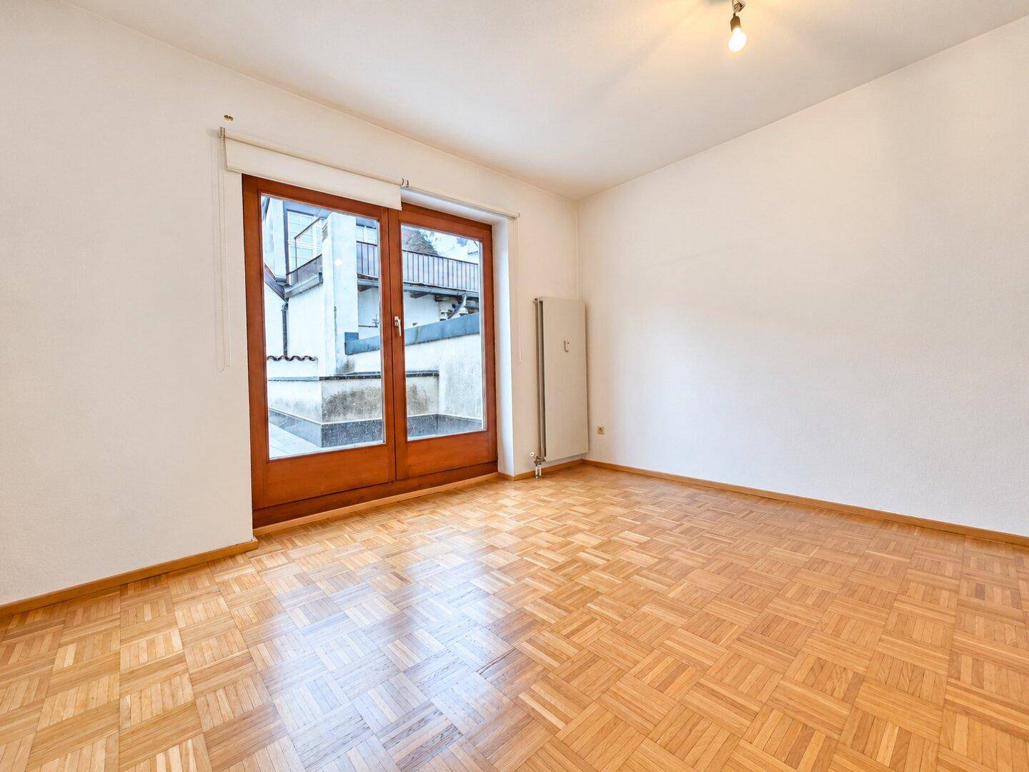 Leeres Zimmer mit Parkettboden und einer großen Schiebetür, die zu einem Außenbereich führt.