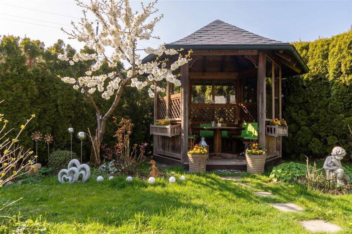 Holzpavillon im grünen Garten, umgeben von blühenden Bäumen und Sträuchern.