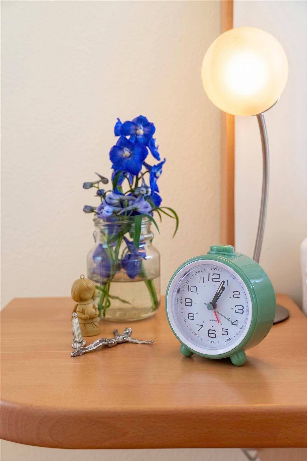 Nachttisch mit Wecker, Lampe und blauen Blumen in einer Vase.