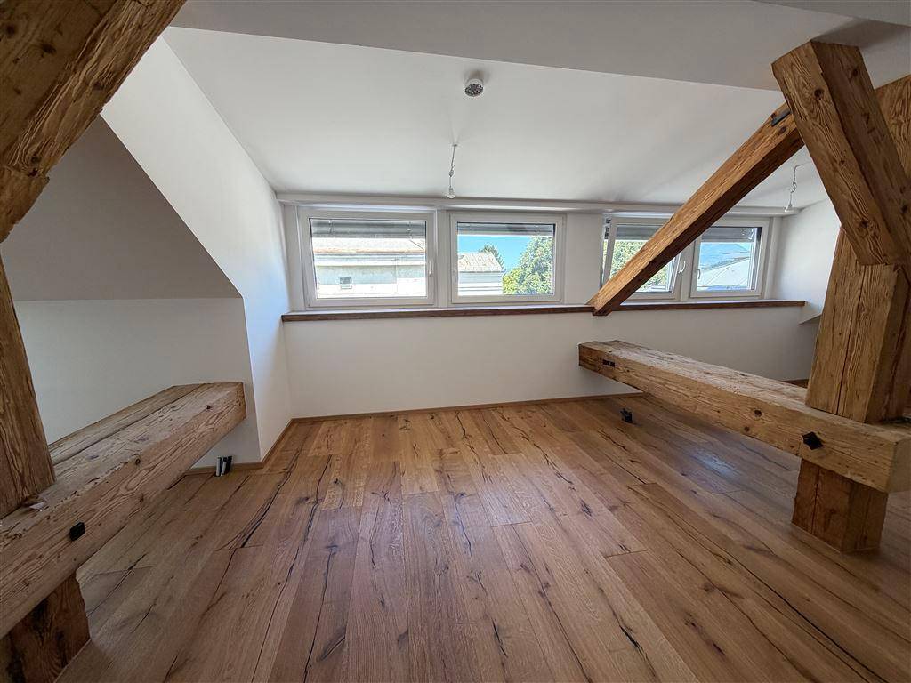 Geräumiger Dachbodenraum mit sichtbaren Holzbalken, Holzboden und einer Reihe von Fenstern für Helligkeit.