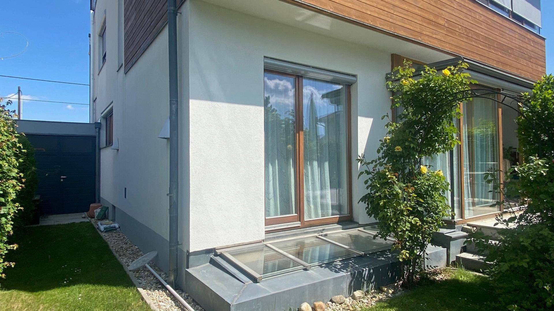 Ansicht der Hausseite mit großen Fenstern, Rollläden und direktem Zugang zum grünen Garten.