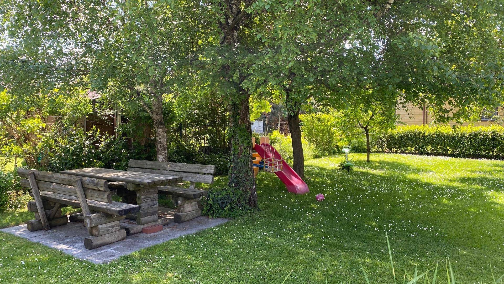 Idyllischer Gartenbereich mit rustikaler Holzbankgarnitur und Kinderrutsche unter Bäumen.