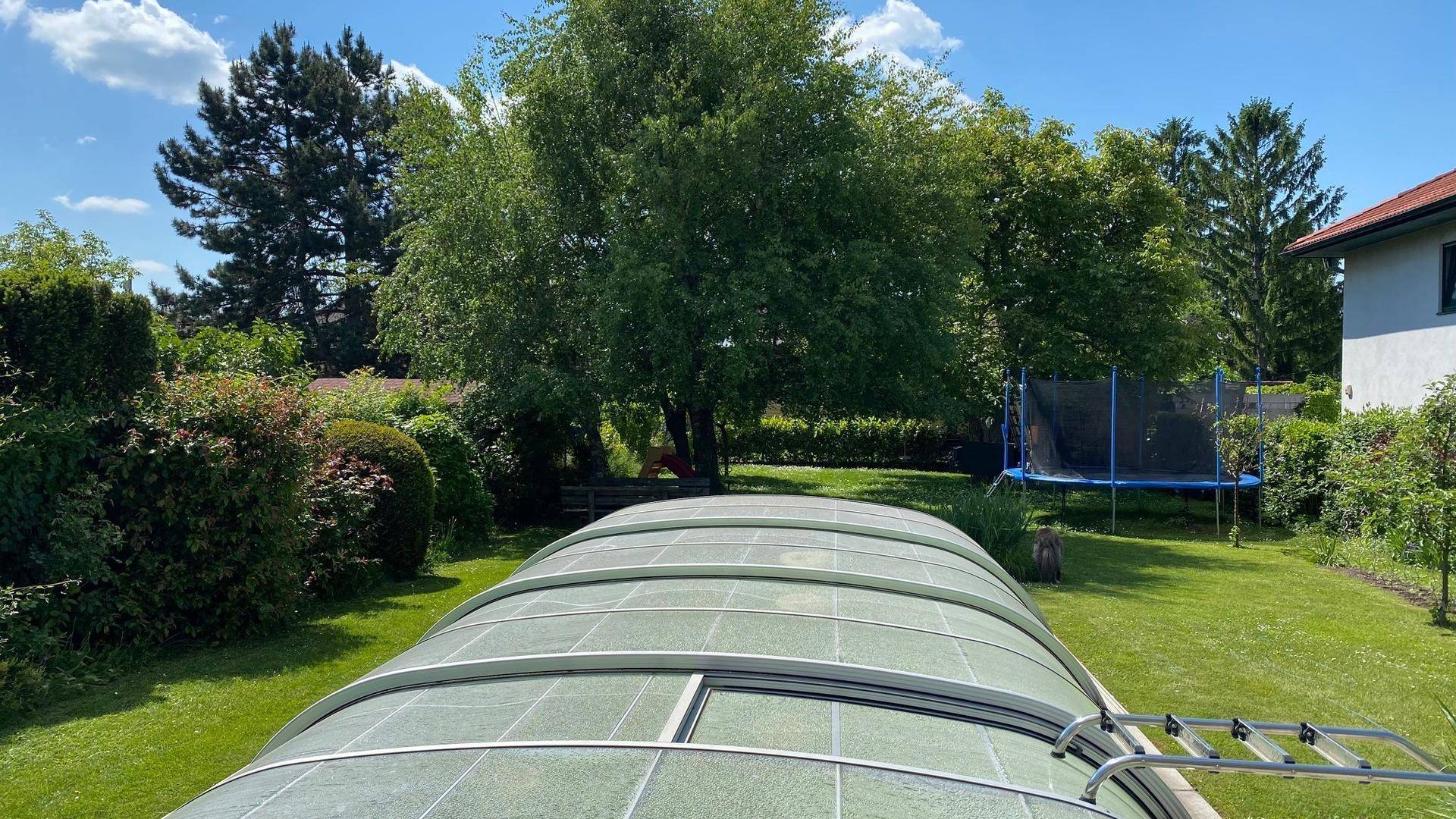 Gepflegter Garten mit großem überdachtem Pool und einem Trampolin im Hintergrund.