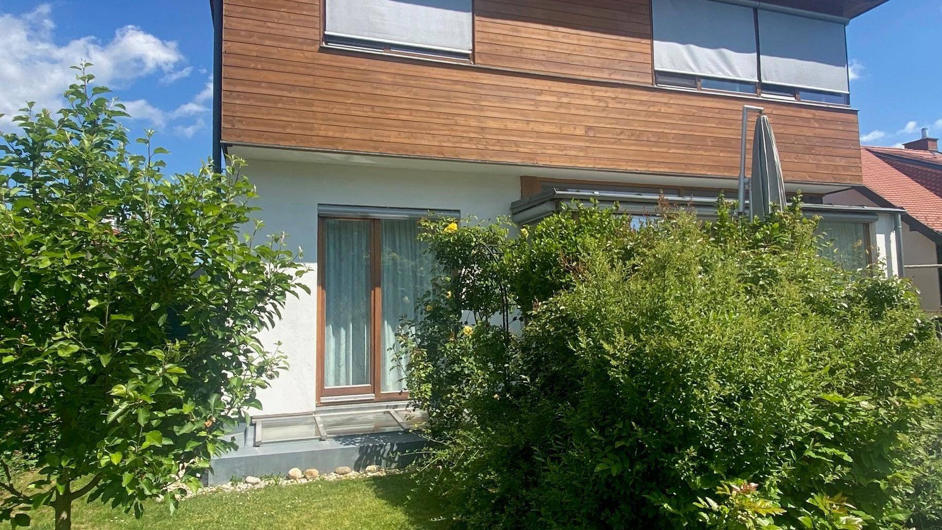 Seitenansicht des Hauses mit Holzfassade, großen Fenstern und üppiger Bepflanzung.