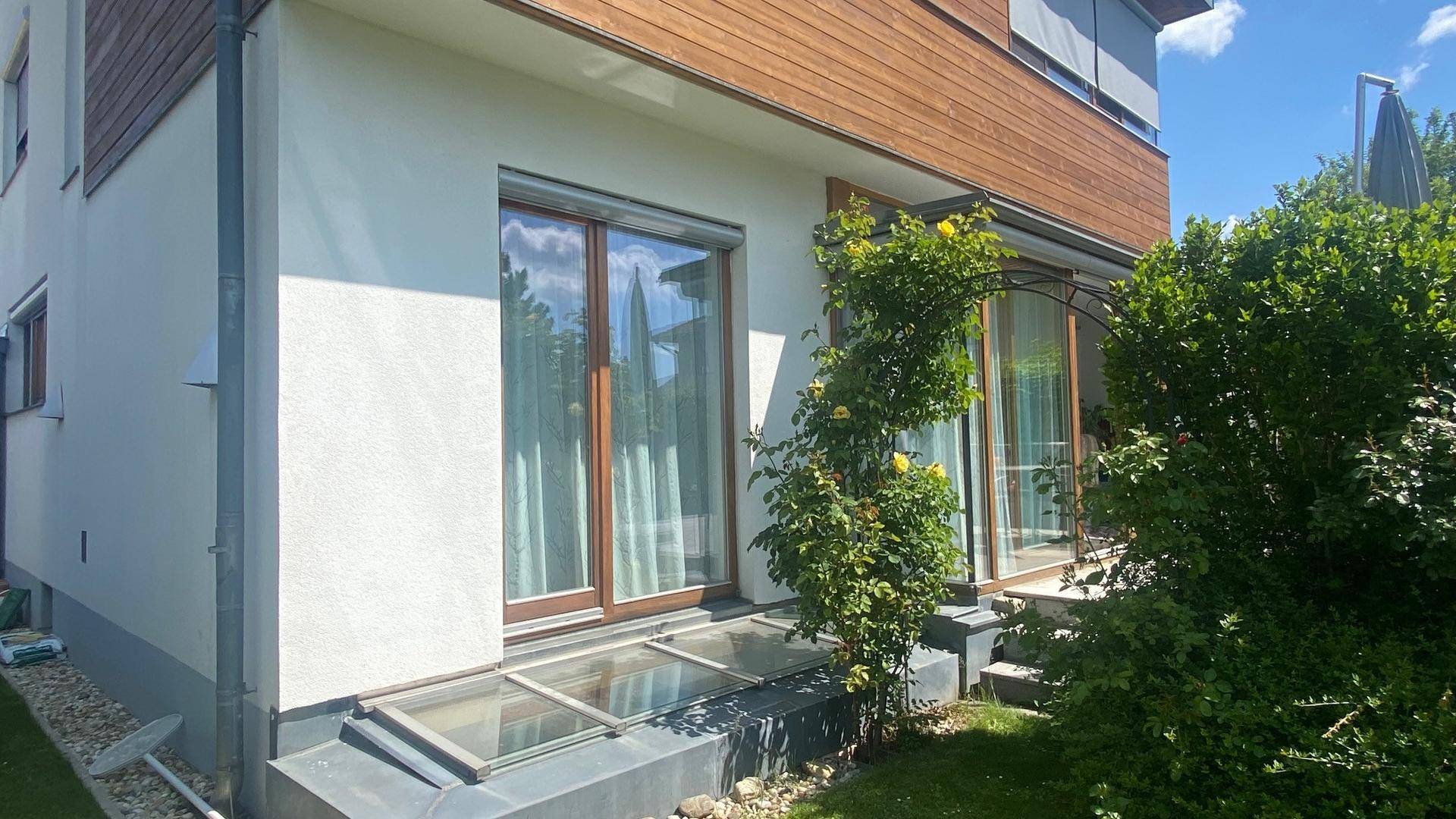 Seitenfassade des Hauses mit großen Fenstern, Rollläden und Blick auf den gepflegten Garten.
