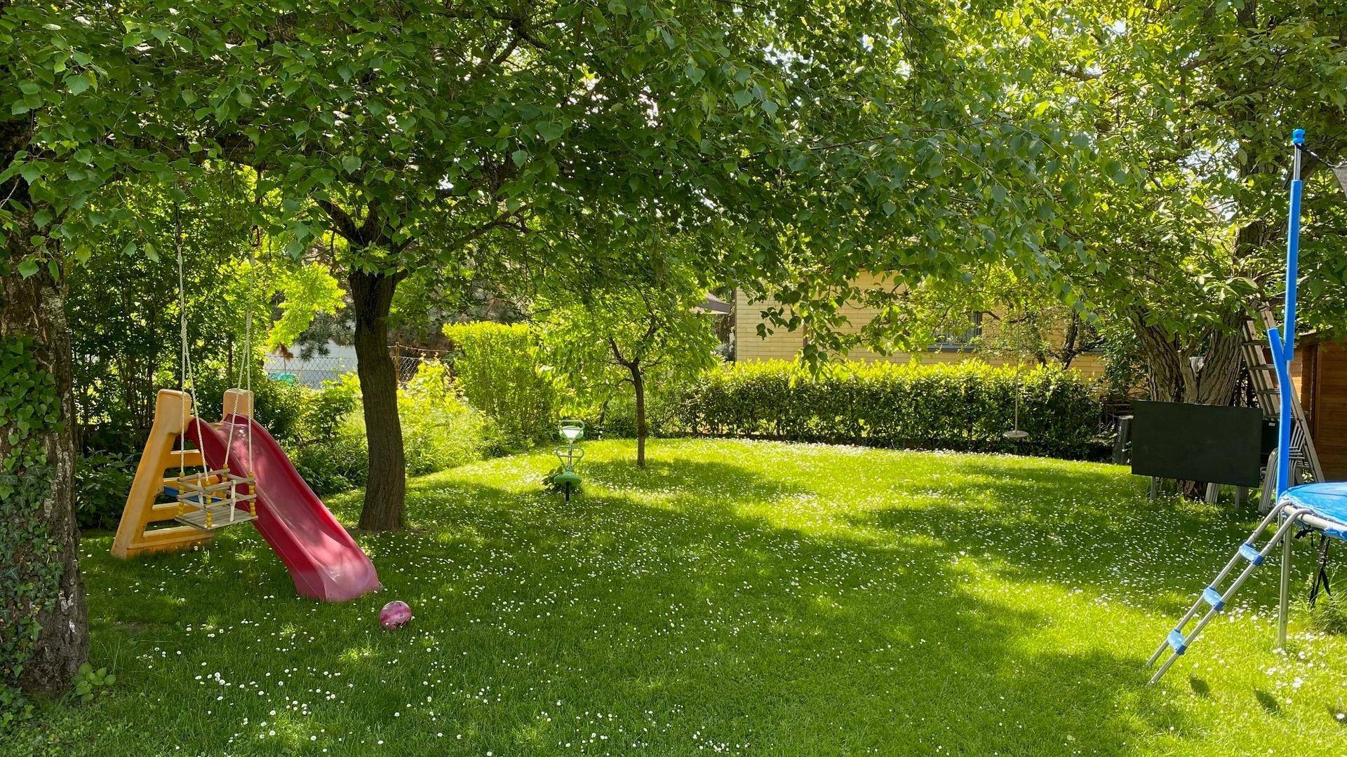 Großer, sonniger Garten mit Rutsche, Schaukel und Trampolin unter schattenspendenden Bäumen.