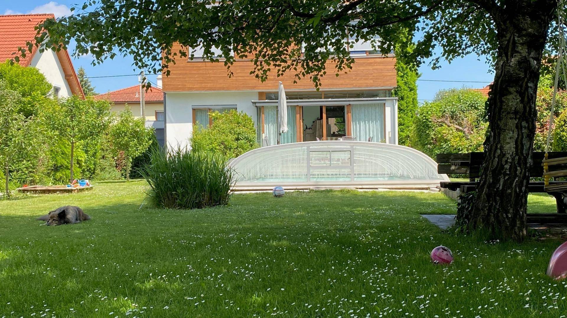 Weitläufiger, sonniger Garten mit überdachtem Pool und Blick auf das moderne Wohnhaus.