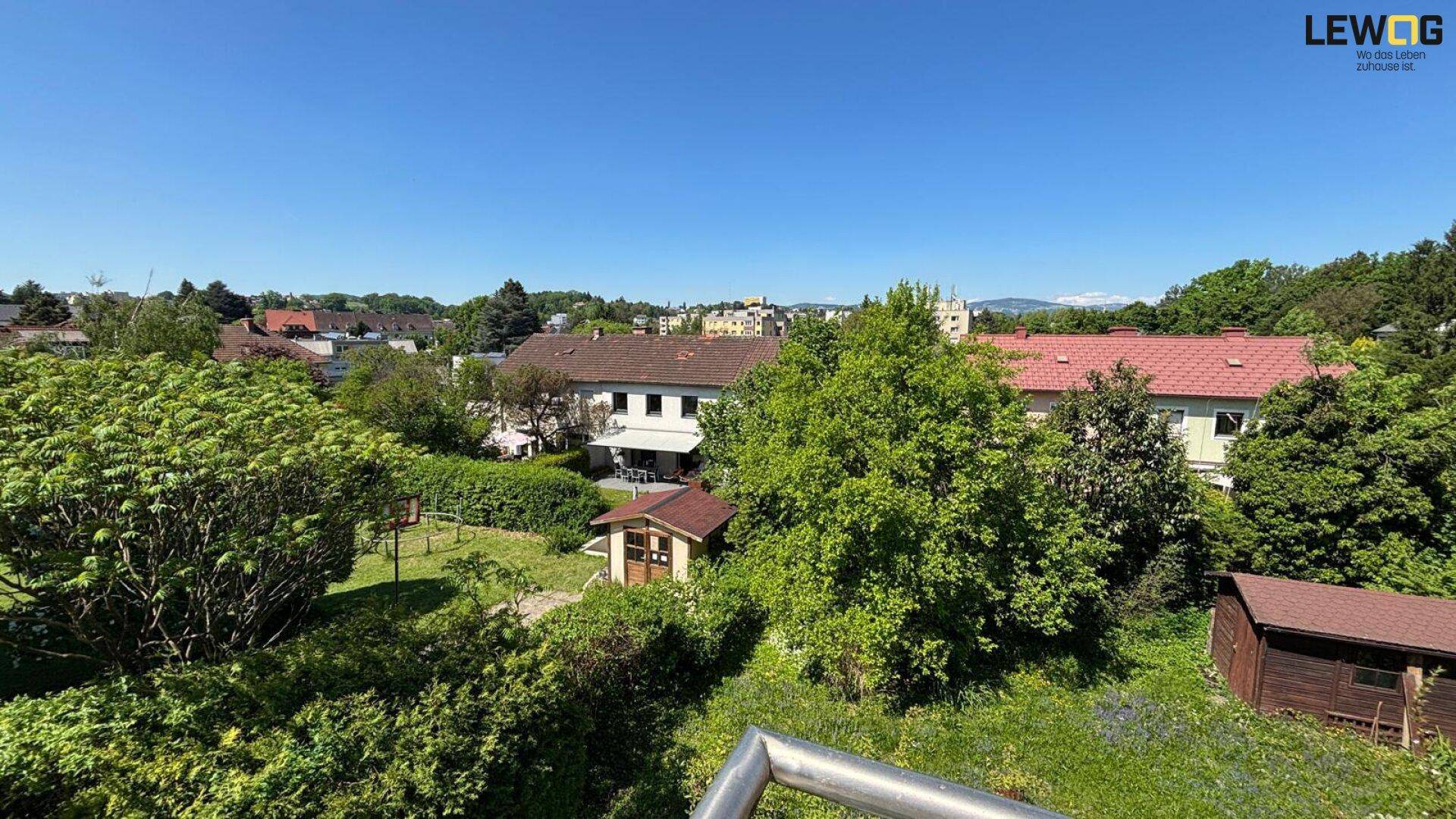 Weitläufiger Blick über die grüne Umgebung mit Wohnhäusern und Bäumen unter blauem Himmel.