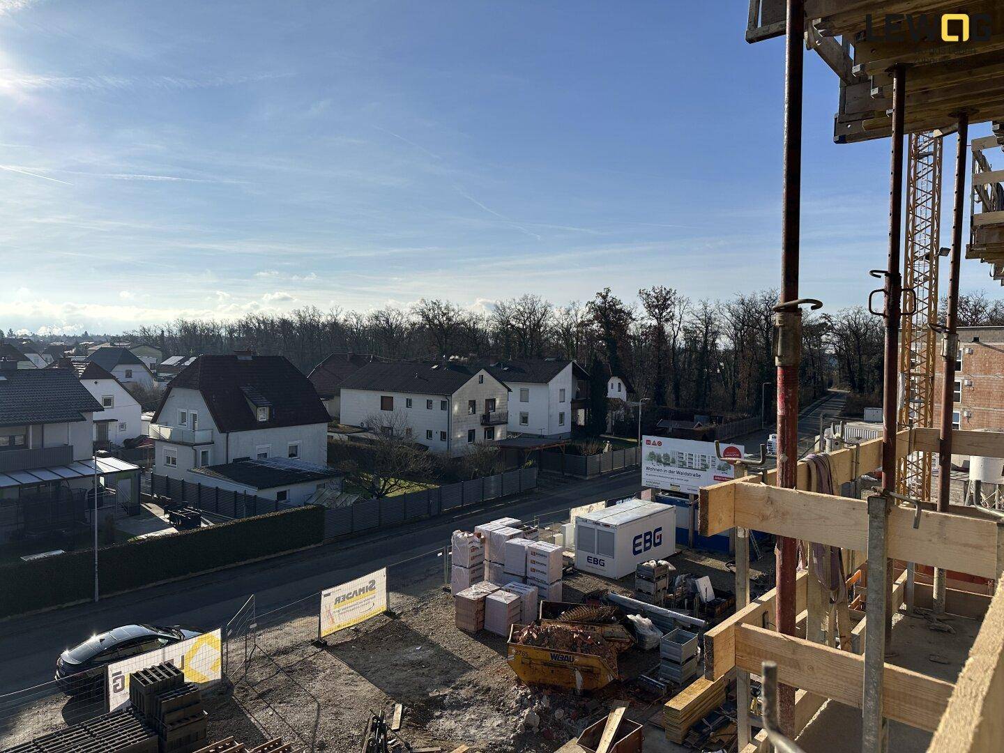 Blick von der Baustelle auf die umliegende Wohnsiedlung und grüne Baumreihen am Horizont.