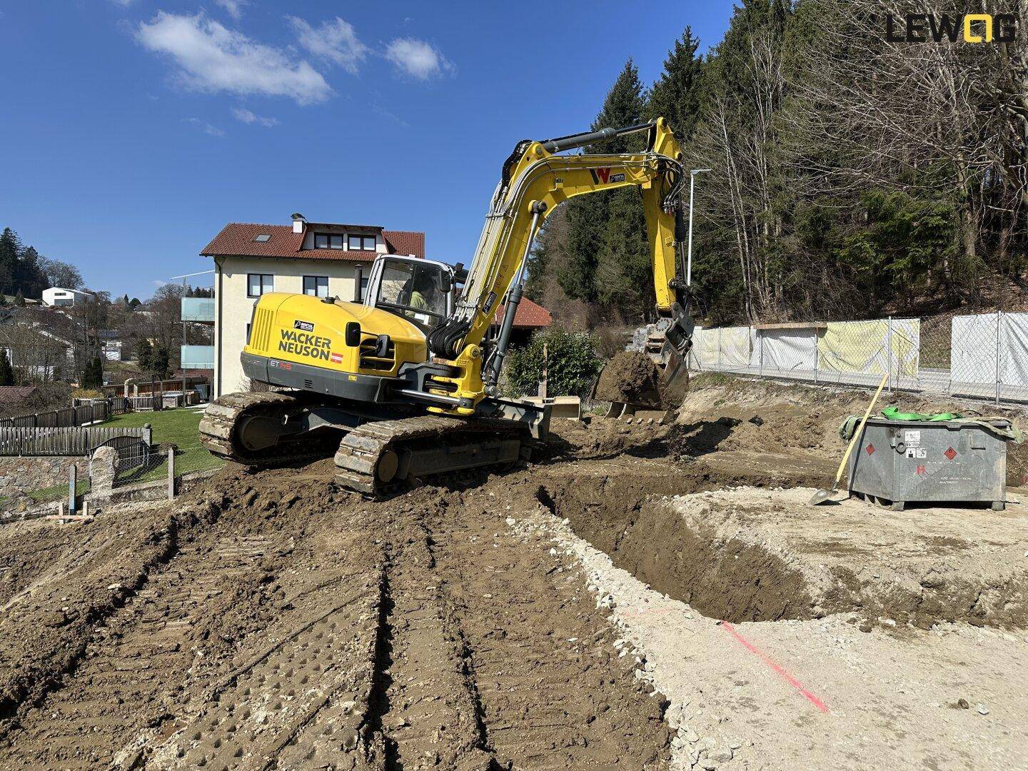 Bagger bei Erdarbeiten auf einer Baustelle mit Wohnhaus und Wald im Hintergrund.