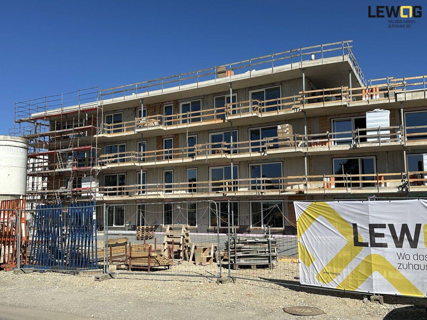 Mehrstöckiges Gebäude im Bau mit Gerüst, großen Fenstern und Bauschild auf der Baustelle.