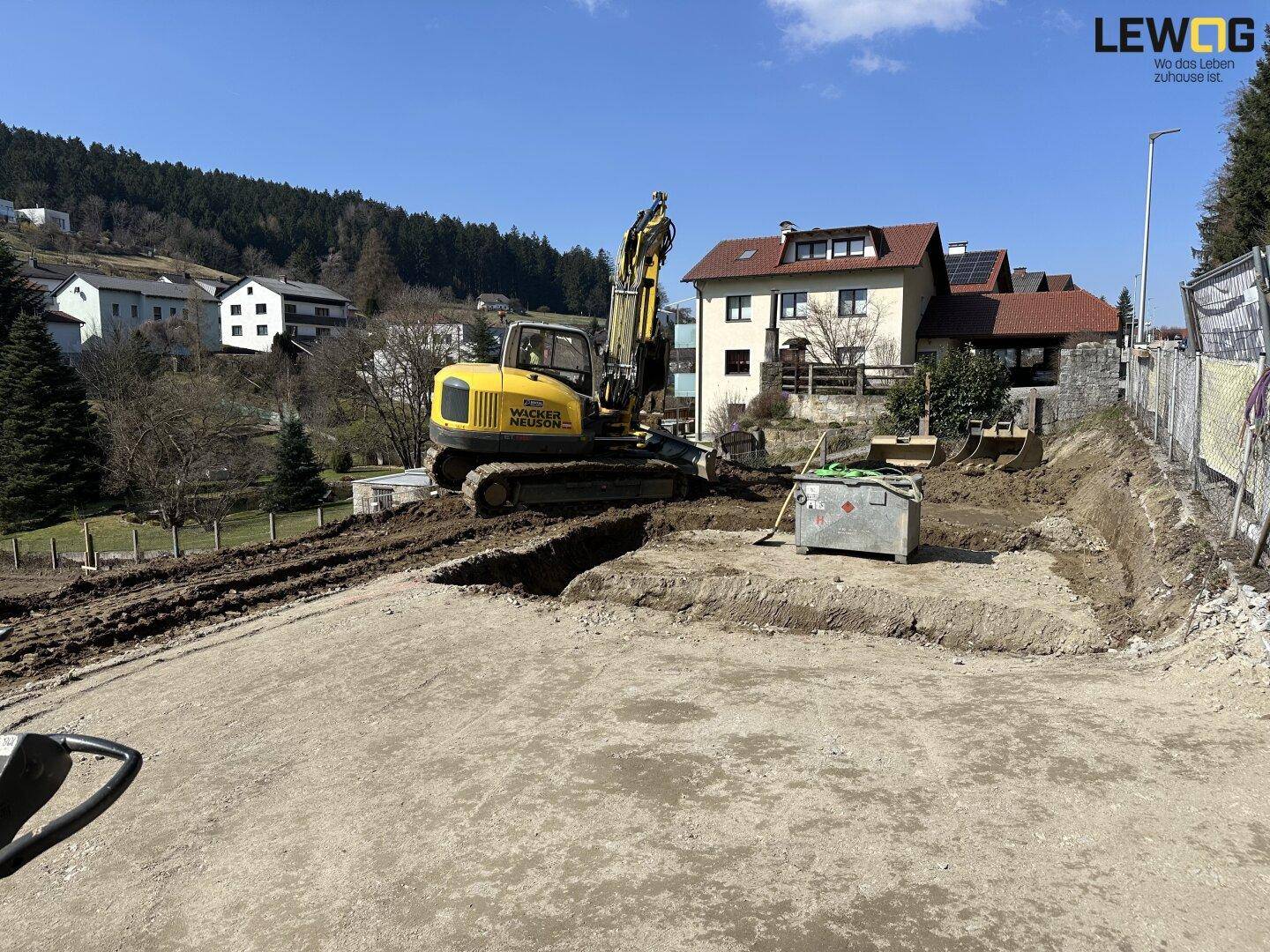 Baustelle mit Bagger und vorbereitetem Baugrundstück, umgeben von Wohnhäusern und einem bewaldeten Hügel.