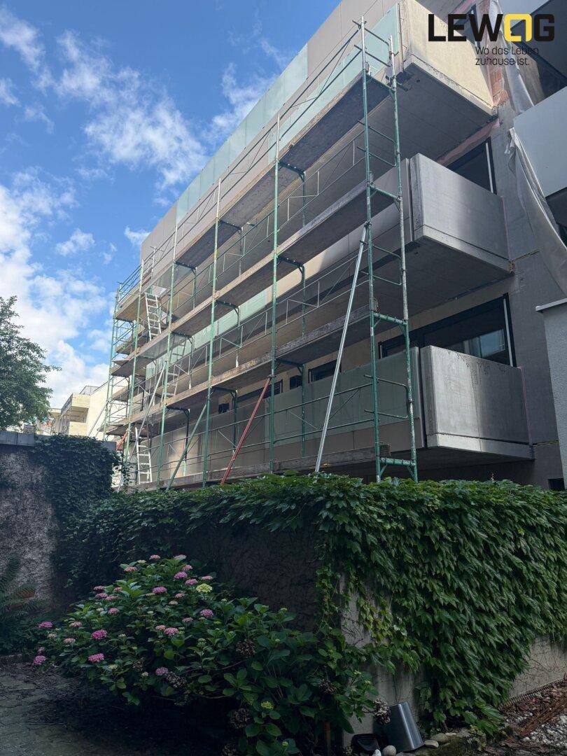 Gebäude im Bau mit Gerüst und Balkonen, umgeben von üppigem Grün und blühenden Pflanzen im Vordergrund.