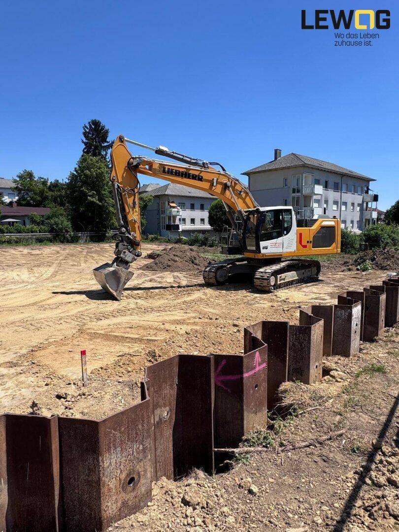 Bagger auf einer Baustelle mit Erdarbeiten und Spundwänden im Vordergrund, umgeben von Wohngebäuden.