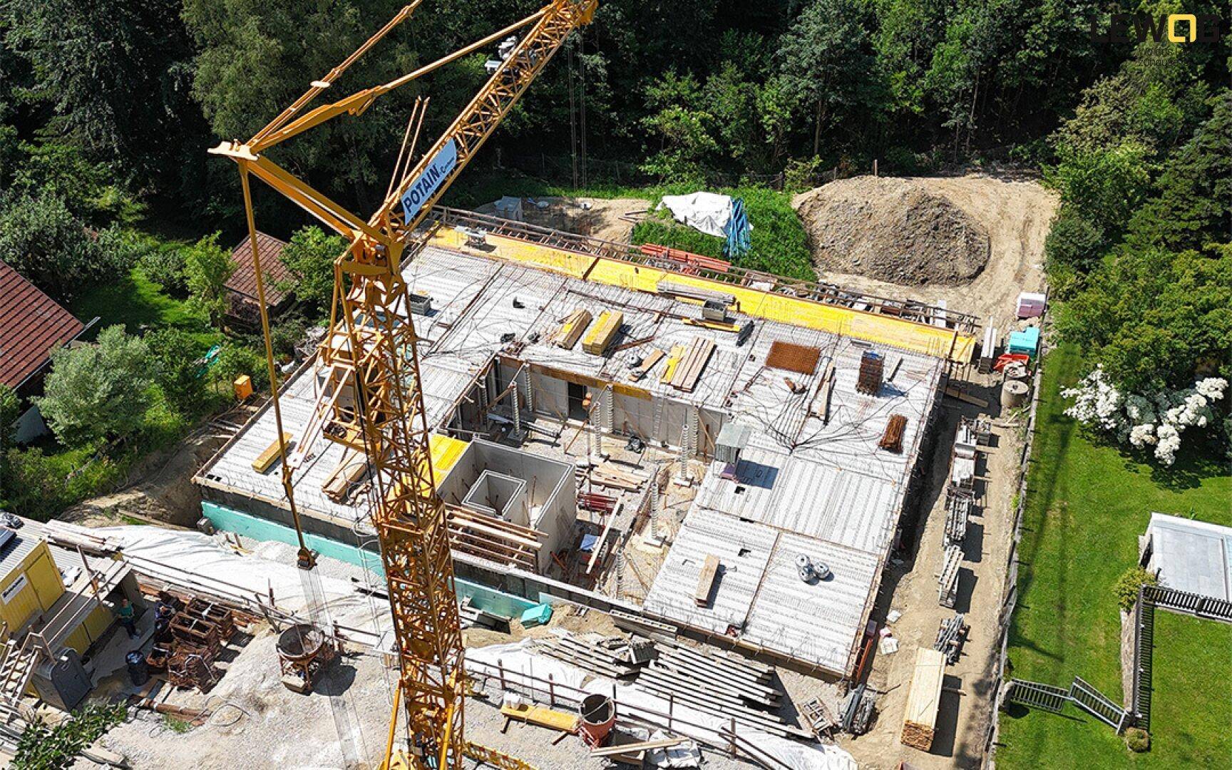Luftaufnahme einer Baustelle mit einem Kran und dem Rohbau eines neuen Gebäudes, umgeben von viel Grün.
