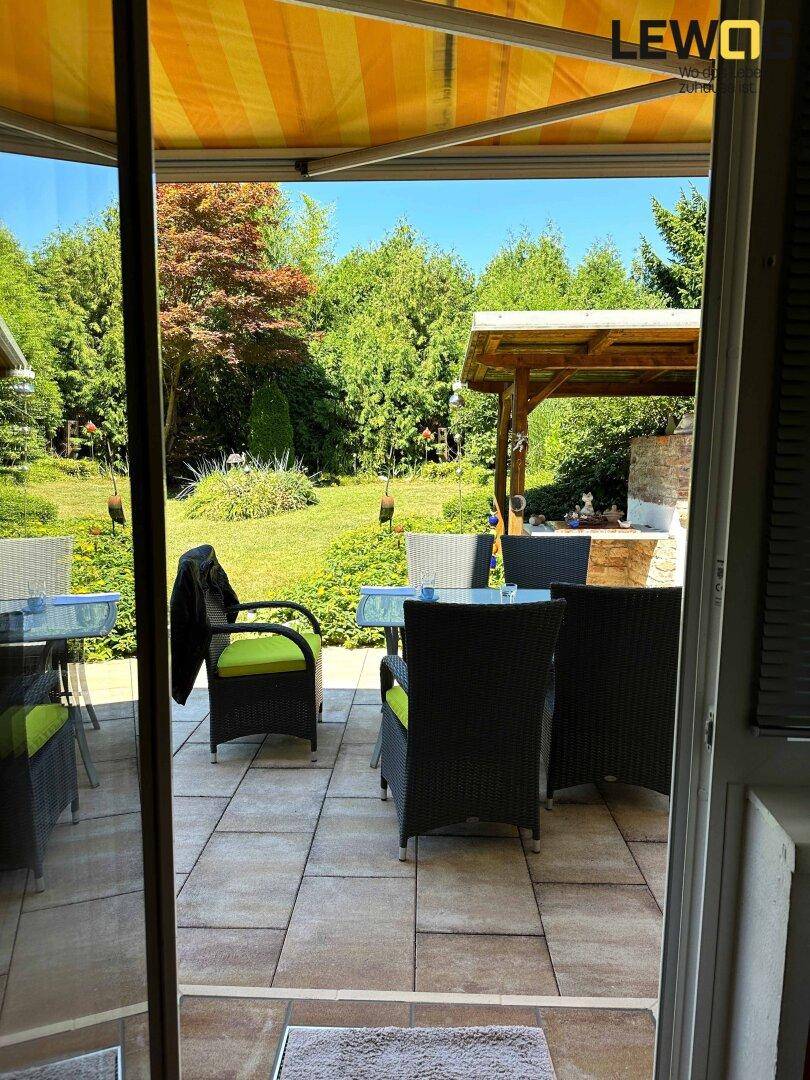 Blick von innen auf die sonnige Terrasse mit Gartenmöbeln und Markise, umgeben von viel Grün.