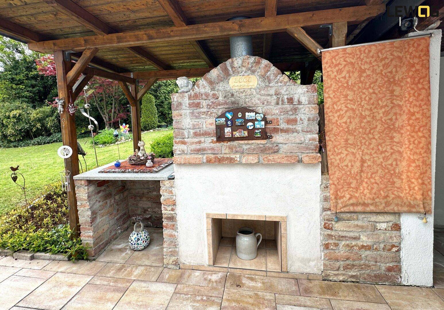 Rustikale Außenküche mit gemauertem Grill und Pizzaofen unter einer Holzpergola im Gartenbereich.