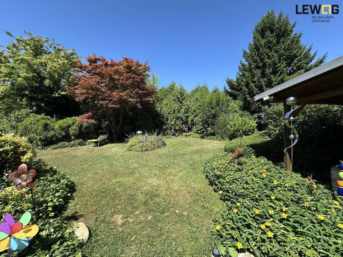Weitläufiger Garten mit üppiger Vegetation, Rasenfläche und einem kleinen Gartenhaus unter blauem Himmel.