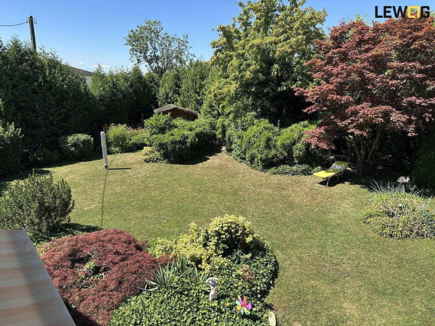 Großer, sonniger Garten mit Rasenfläche, vielfältiger Bepflanzung und einem Gartenhaus im Hintergrund.