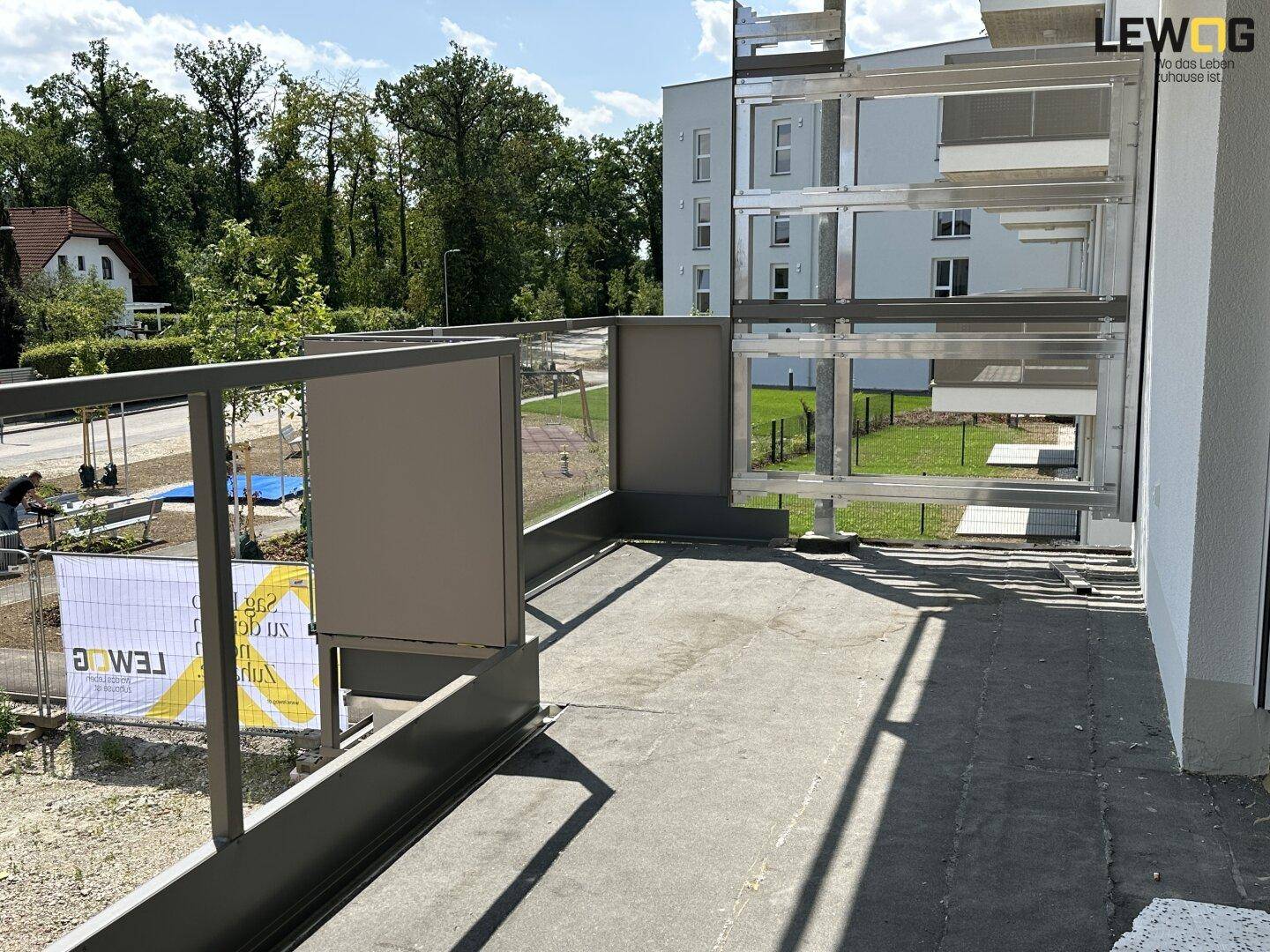 Unfertiger Balkon mit Metallgeländer und Blick ins Grüne sowie auf ein weiteres Gebäude.