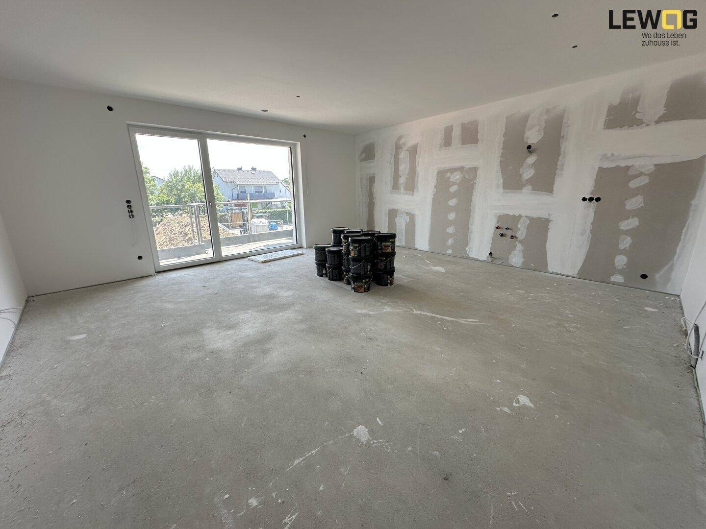 Geräumiger, unfertiger Wohnraum mit Betonboden und großem Schiebefenster zum Außenbereich.