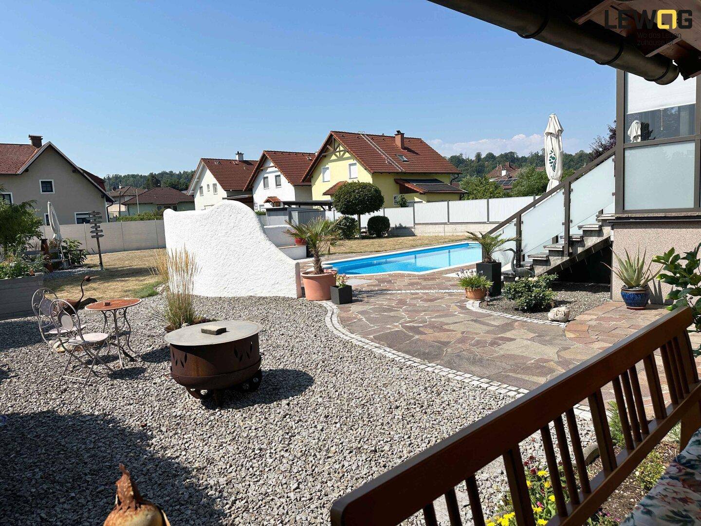 Kiesgarten mit Feuerstelle, gepflastertem Weg und Pool im Hintergrund unter blauem Himmel.