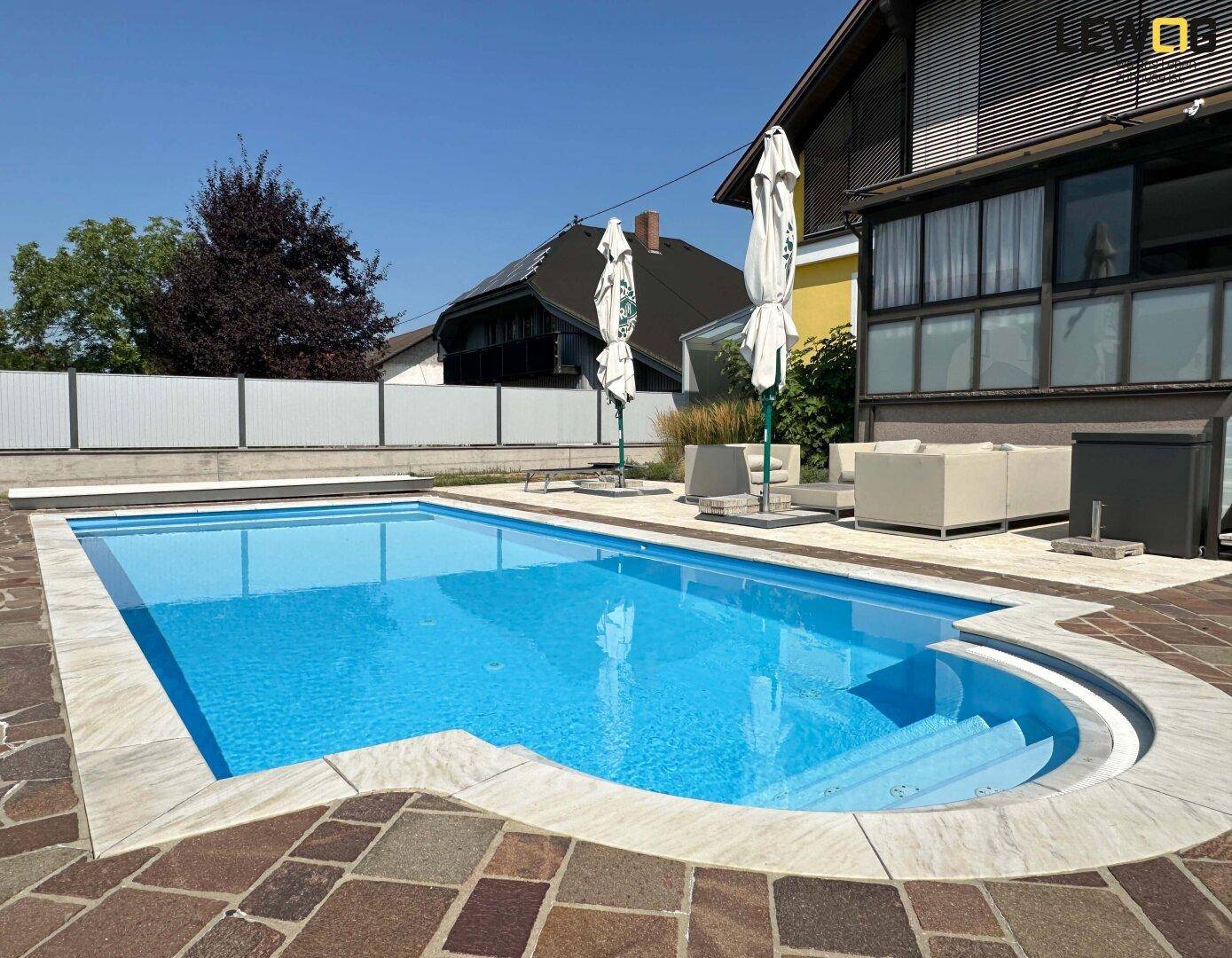 Blauer Swimmingpool mit Steinfliesenrand und gepflasterter Terrasse im sonnigen Garten.