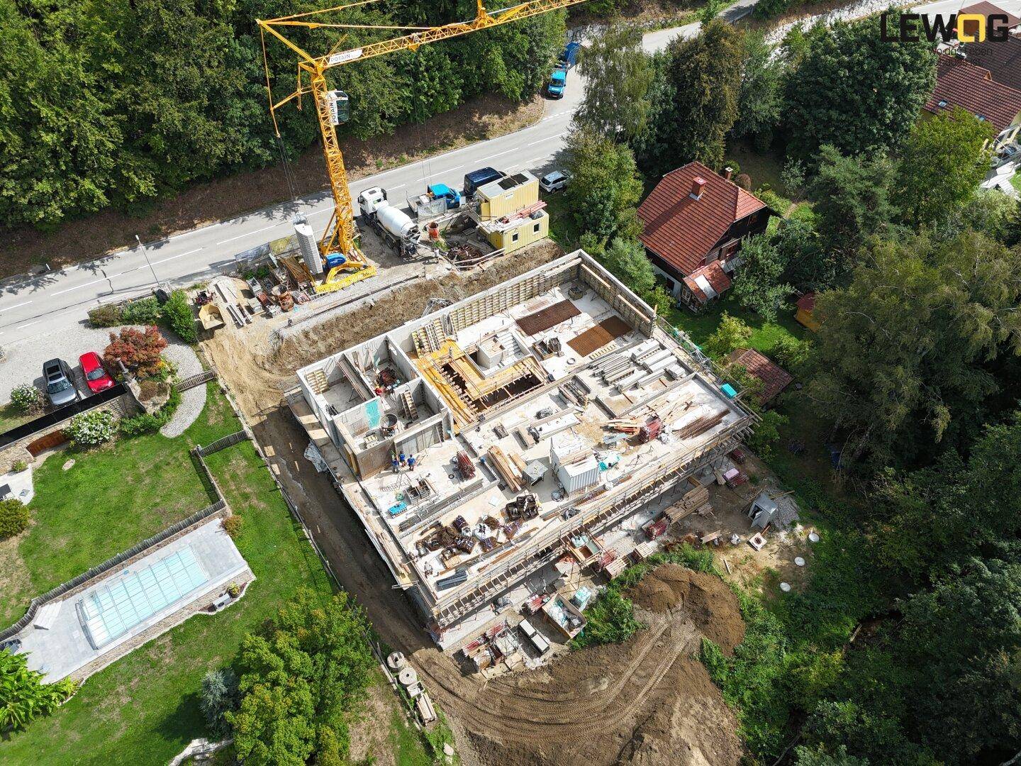Luftaufnahme einer Baustelle für ein neues Gebäude mit Kran, umgeben von viel Grün und einem Pool.