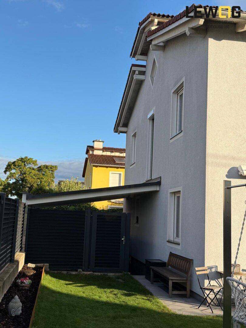 Seitlicher Gartenbereich mit Rasen, Sichtschutzzaun und überdachtem Zugang zum Haus unter blauem Himmel.