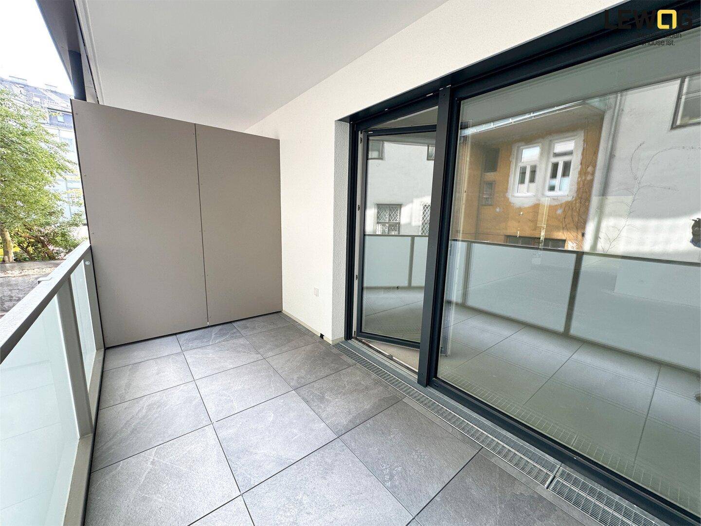 Moderner Balkon mit grauen Fliesen, Glasbrüstung und großer Schiebetür zum Innenraum.