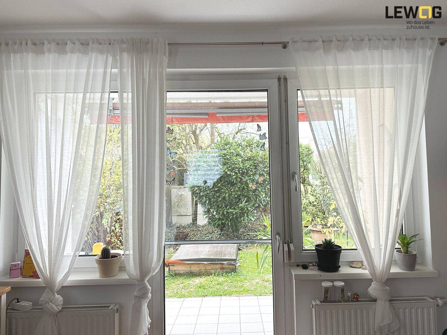 Heller Innenraum mit großen Fenstern und Zugang zum Garten, geschmückt mit weißen Vorhängen.