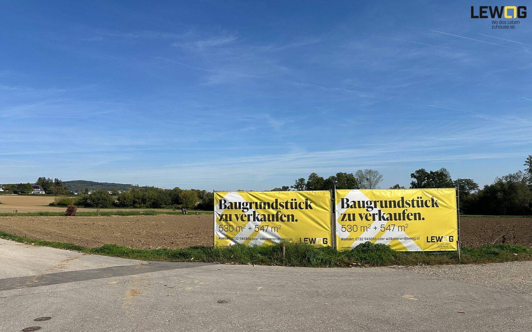 Zwei gelbe Banner bewerben ein Baugrundstück zum Verkauf in ländlicher Umgebung unter blauem Himmel.