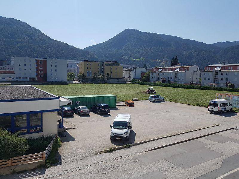 Außenansicht des Gewerbegebäudes mit Parkplatz, umgeben von Grünflächen und Wohngebäuden vor einer Bergkette.