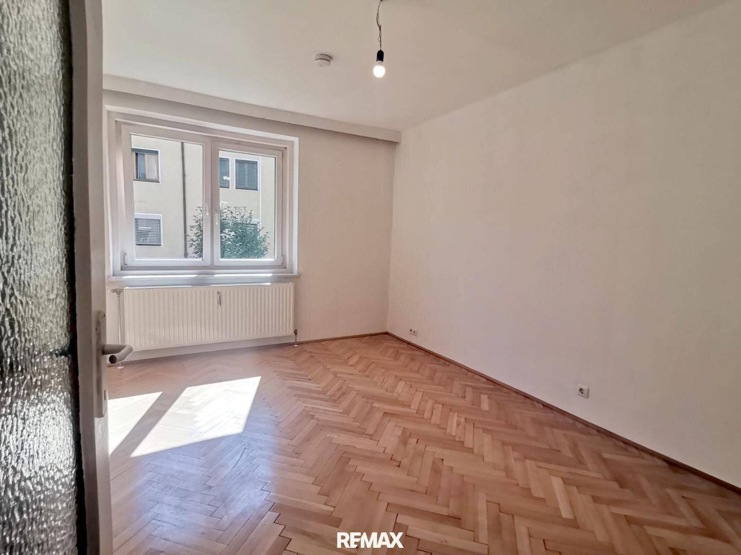 Heller Raum mit Fischgrätparkettboden und großem Fenster, das viel Tageslicht hereinlässt.