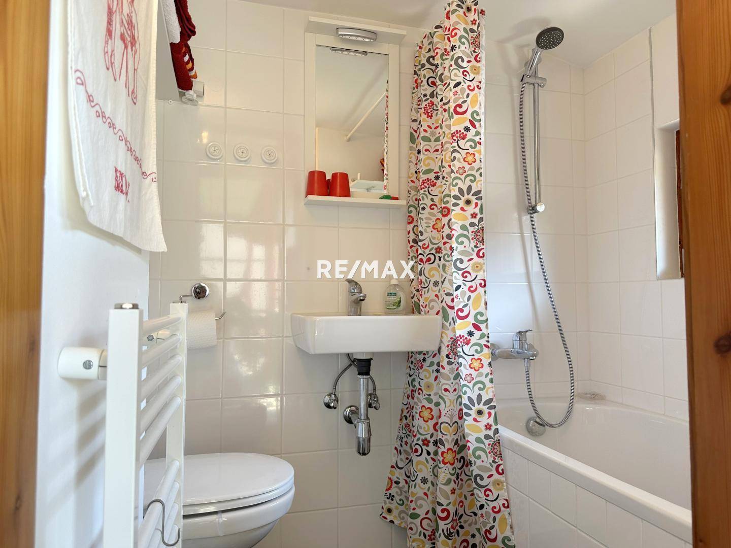 Kleines Badezimmer mit Badewanne, Duschvorhang und beheiztem Handtuchhalter, weiß gefliest.