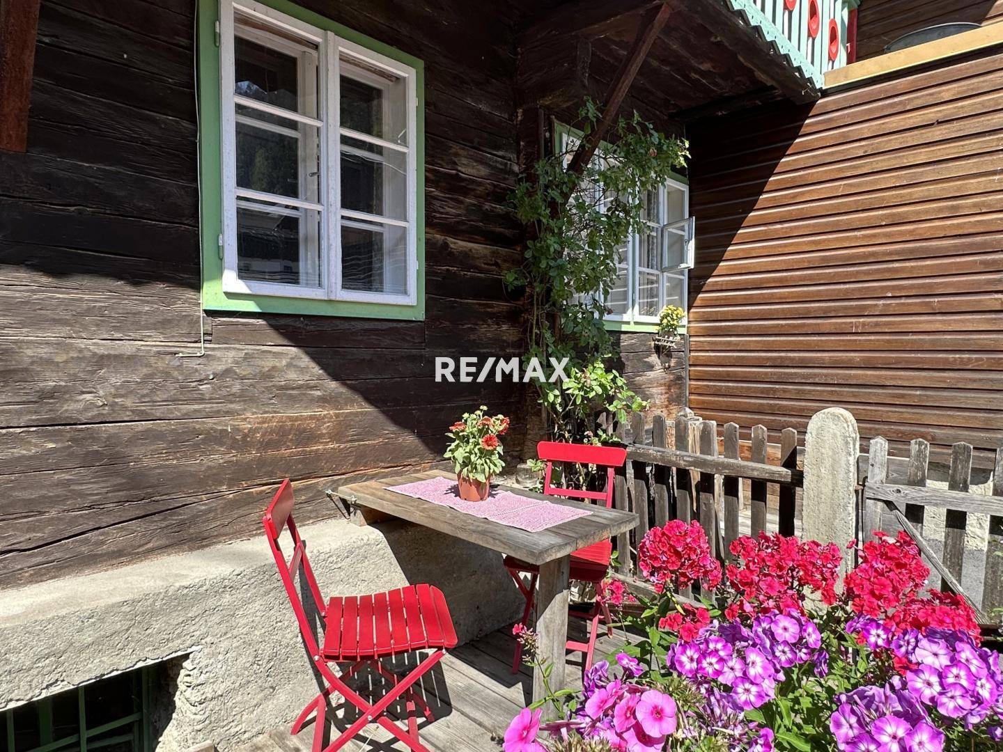 Gemütlicher kleiner Sitzbereich im Freien mit Holztisch und roten Stühlen, umgeben von Blumen.