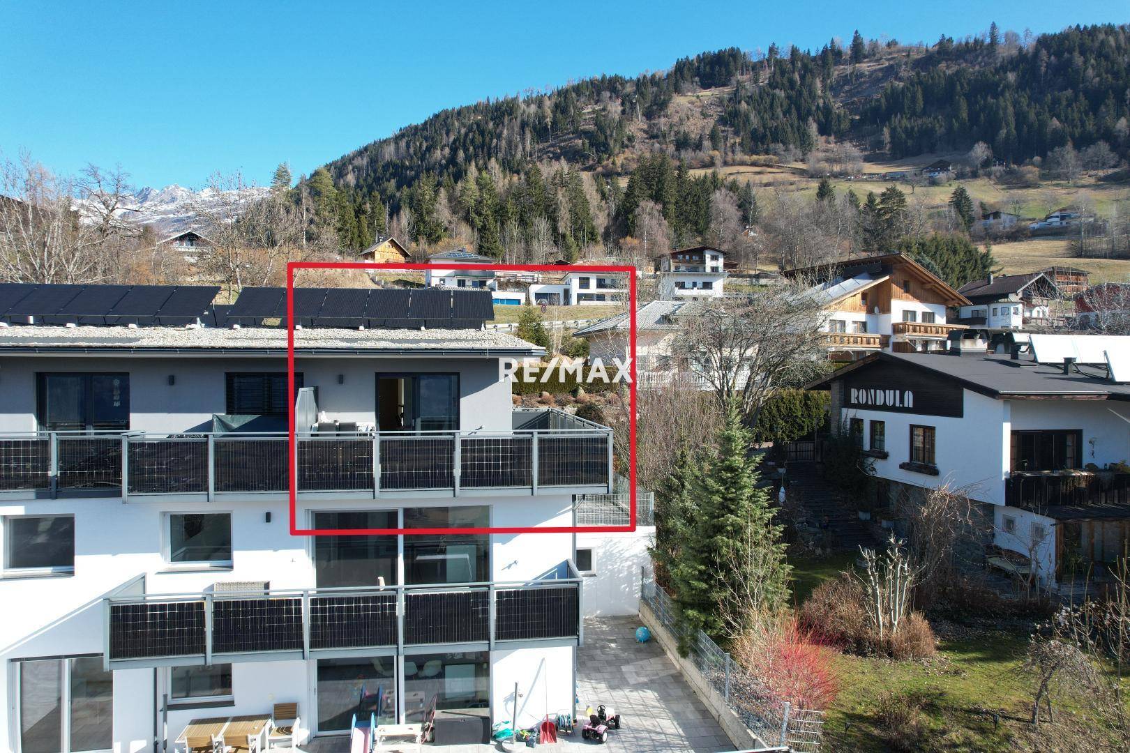 Luftaufnahme eines modernen Mehrfamilienhauses mit Balkonen und Solarmodulen in bergiger Umgebung.
