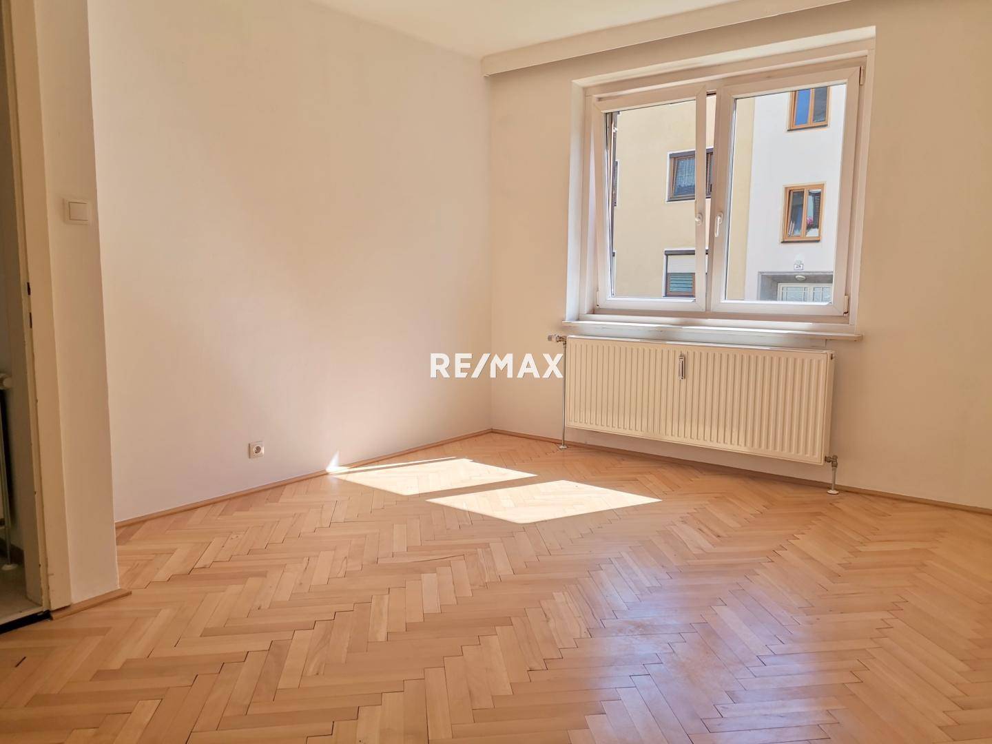 Helles Schlafzimmer mit Fischgrätparkett und großem Fenster, das viel Tageslicht hereinlässt.