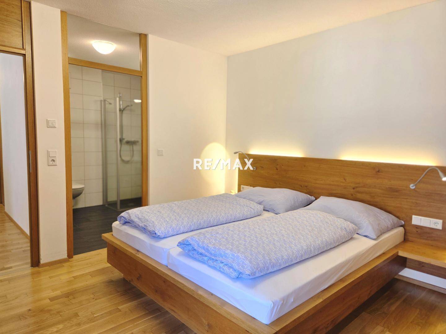 Schlafzimmer mit Doppelbett und Holzkopfteil, direktem Zugang zum Bad und hellem Parkettboden.