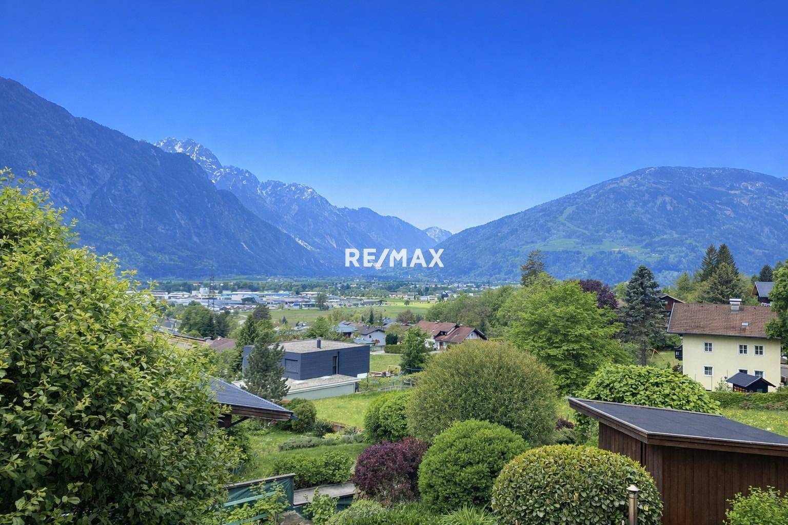 Panoramablick auf eine grüne Tallandschaft mit majestätischen Bergen im Hintergrund und einer Wohnsiedlung.
