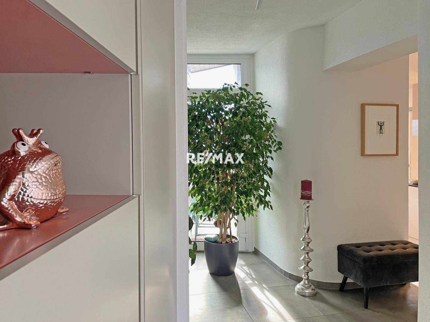 Heller Eingangsbereich mit großem Fenster, dekorativer Pflanze und modernem Hocker.