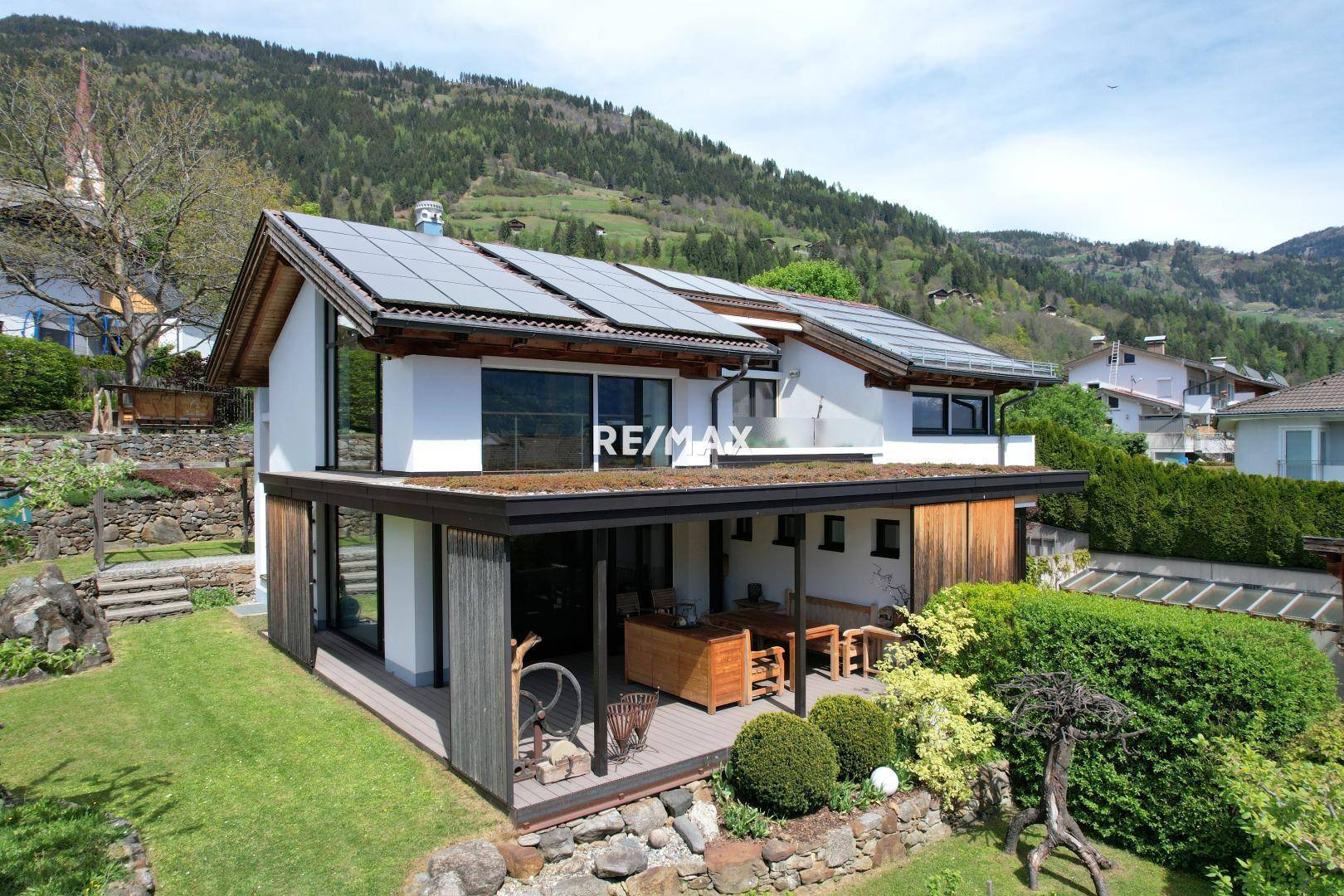 Modernes Haus mit großer überdachter Terrasse, Photovoltaikanlage und Blick auf die umliegende Berglandschaft.