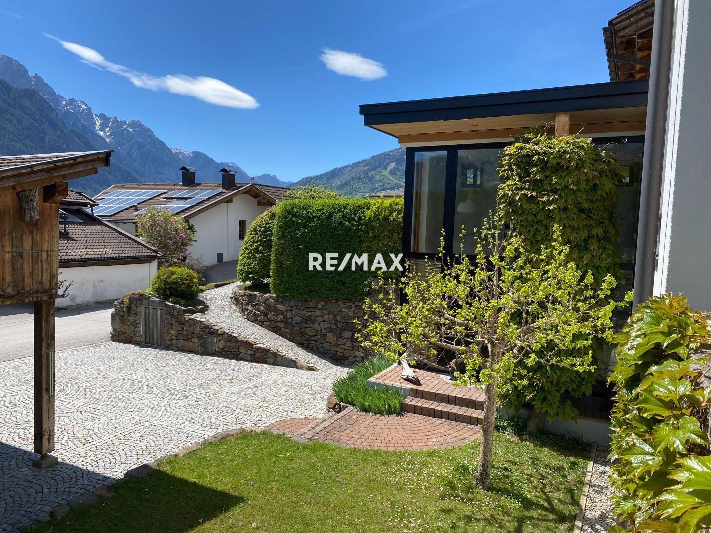 Eingangsbereich des Hauses mit gepflasterter Auffahrt und gepflegter Hecke vor alpiner Kulisse.