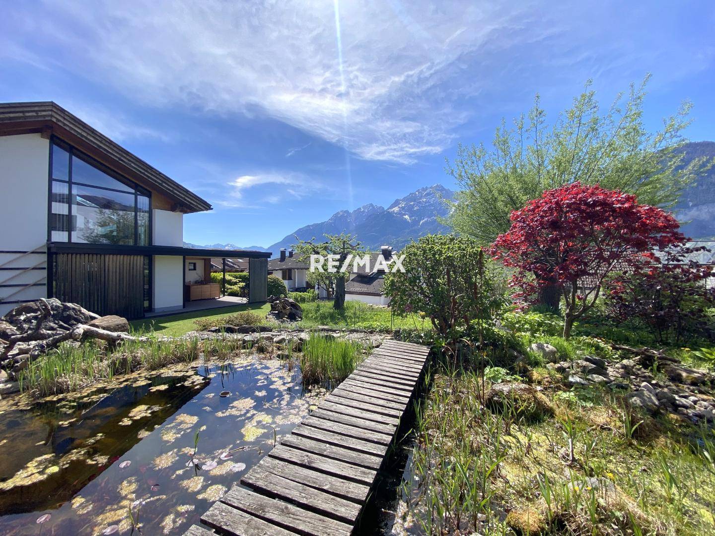 Modernes Haus mit weitläufigem Garten, Teich und Holzsteg vor einer beeindruckenden Bergkulisse.