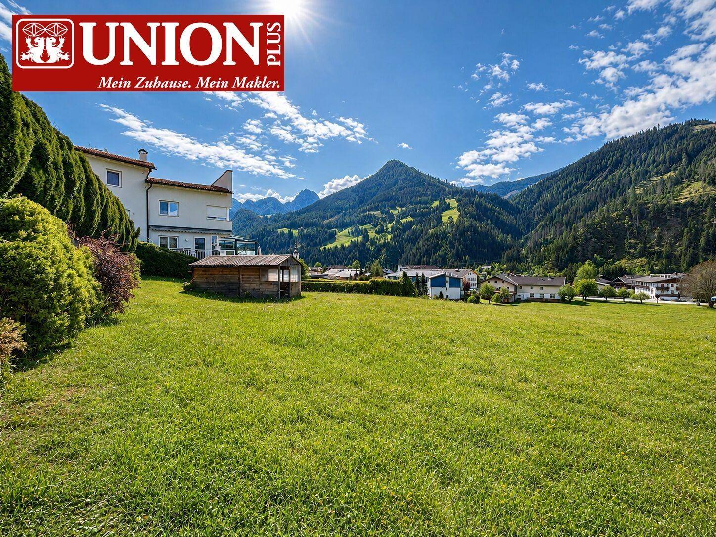 Sonnige Wiesenfläche mit einem Haus im Vordergrund und Panoramablick auf die Alpen.