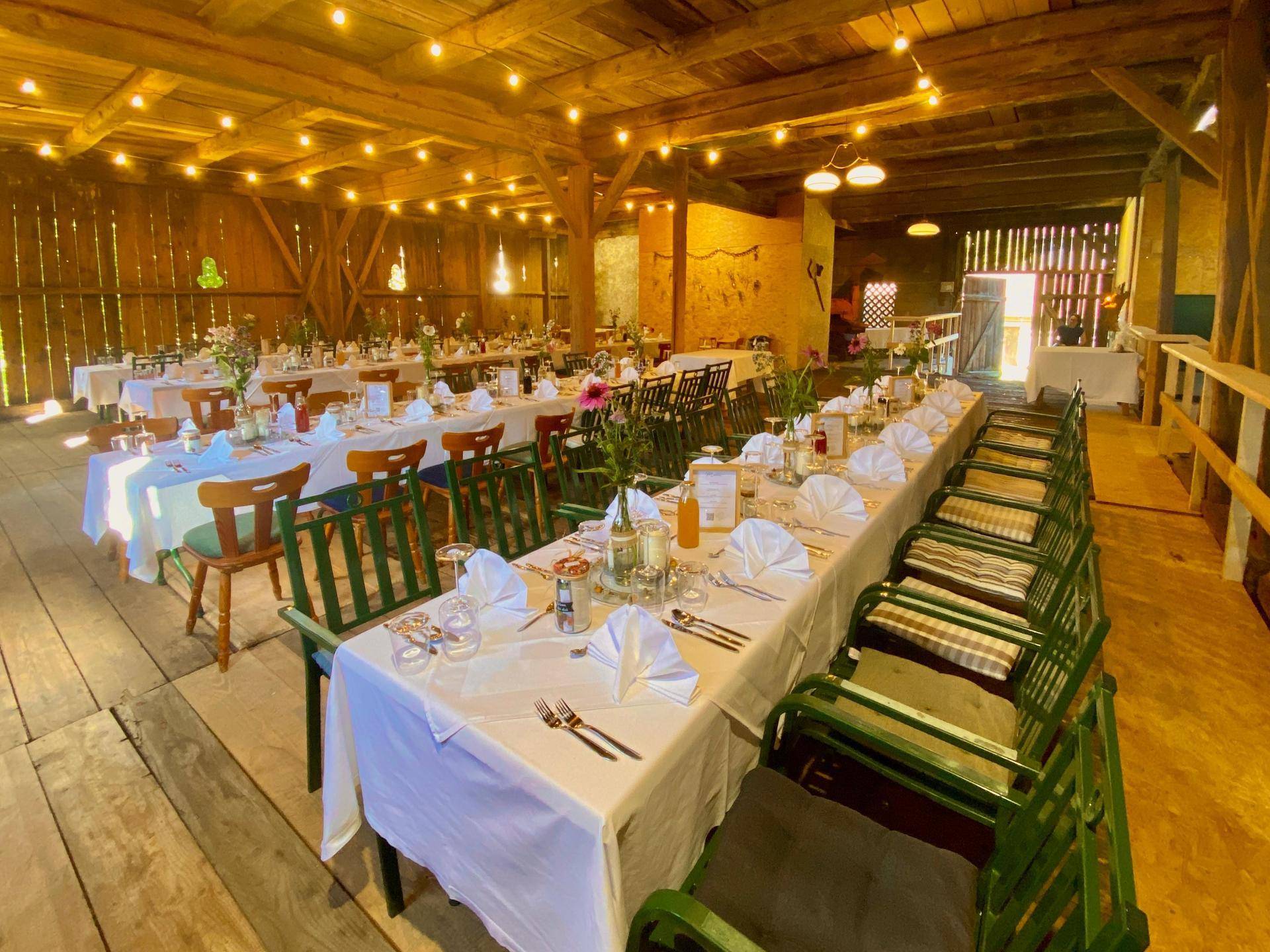 Rustikaler Veranstaltungsraum mit langen Tischen, sichtbaren Holzbalken und festlicher Beleuchtung für Feierlichkeiten.