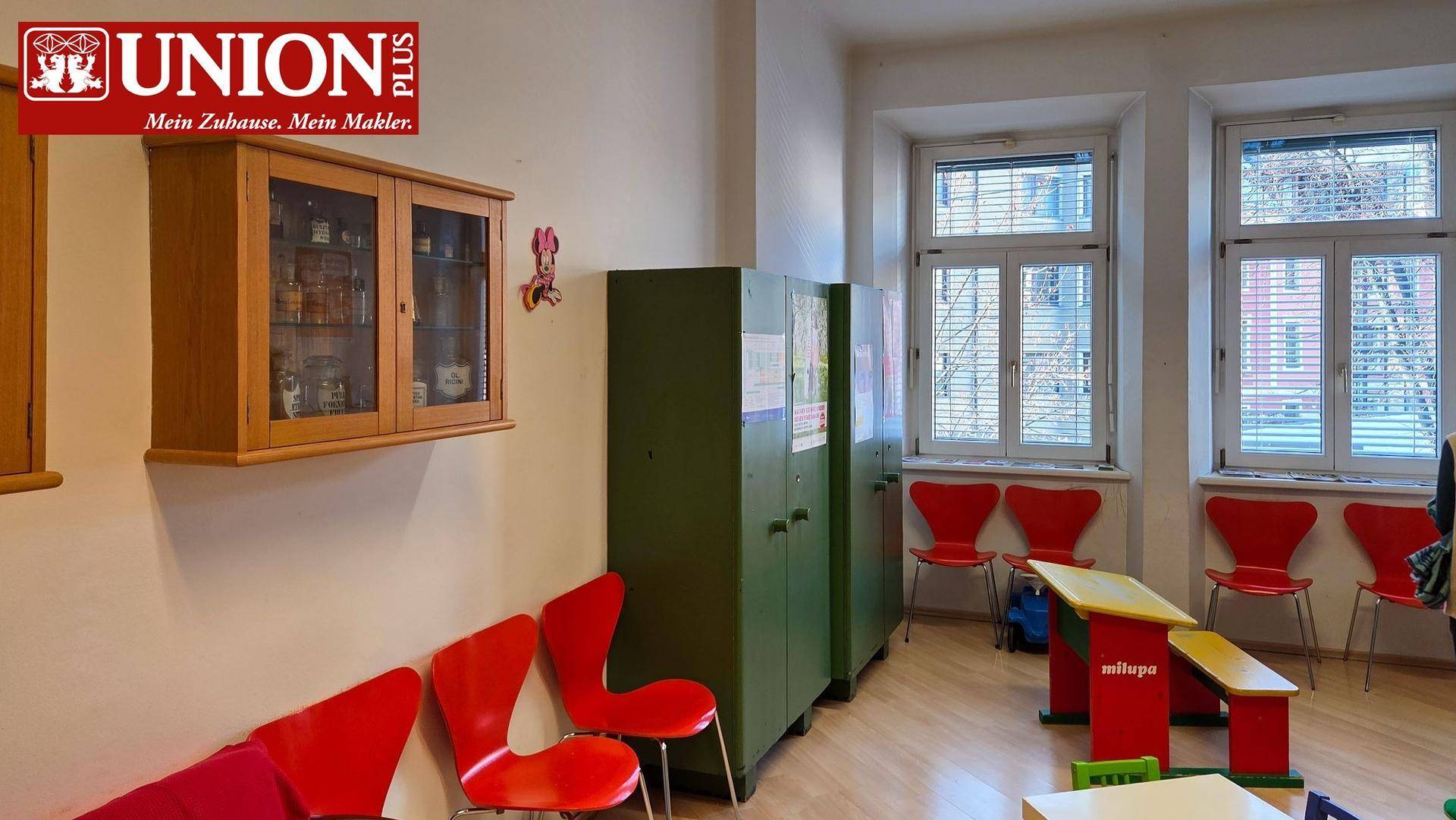 Warteraum oder Spielbereich mit roten Stühlen, grünen Schränken und hellen Fenstern.