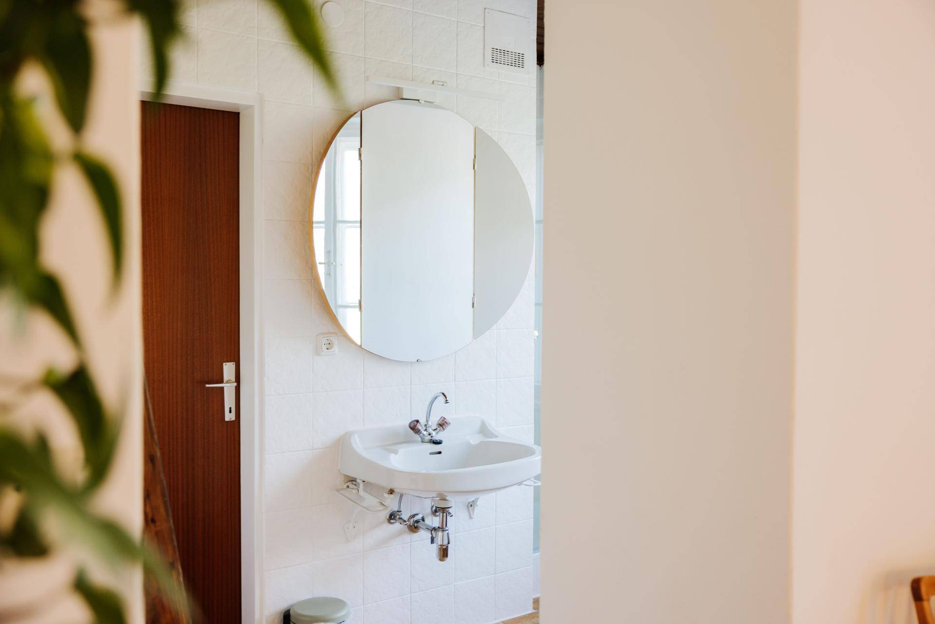 Badezimmer mit weiß gefliesten Wänden, einem runden Spiegel über dem Waschbecken und einem Abluftventilator.